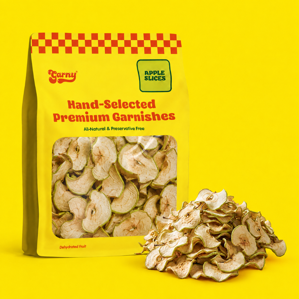 A yellow bag of Garny Hand-Selected Premium Garnishes apple slices stands on a bright yellow background with a pile of dried apple slices beside it. The bag has a red-and-yellow checkered pattern at the top, a clear window showing the slices, and a green label reading Apple Slices.