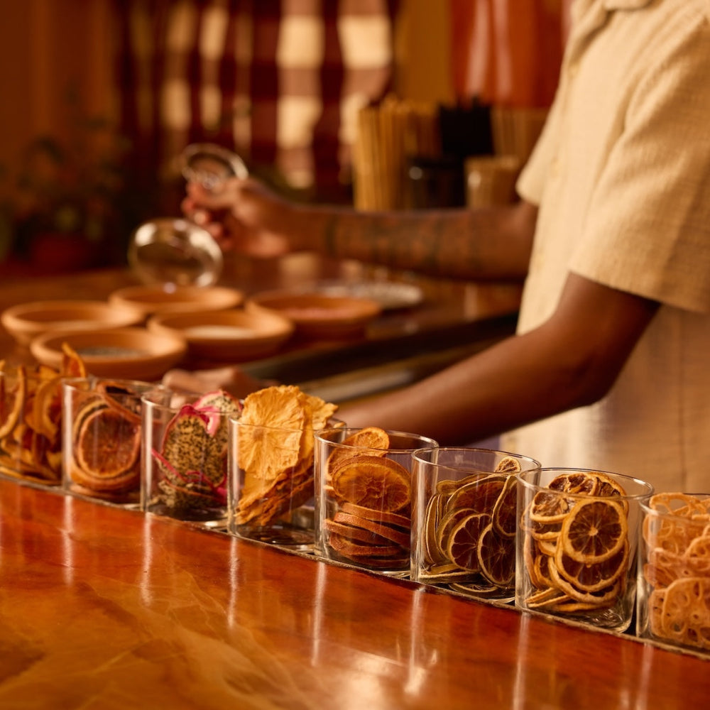 A row of clear glass cups filled with dried citrus slices and dehydrated cocktail garnishes arranged on a polished wooden bar counter, with a bartender in a cream shirt working in the background.