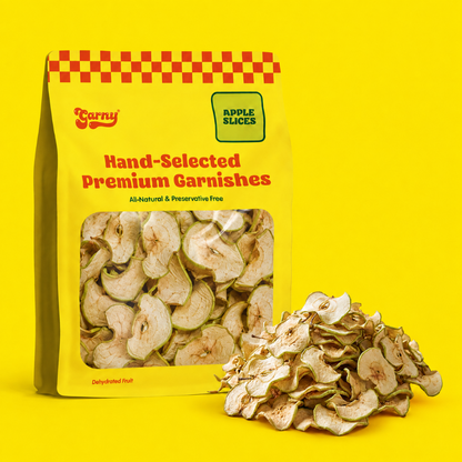 A yellow bag of Garny Hand-Selected Premium Garnishes apple slices stands on a bright yellow background with a pile of dried apple slices beside it. The bag has a red-and-yellow checkered pattern at the top, a clear window showing the slices, and a green label reading Apple Slices.