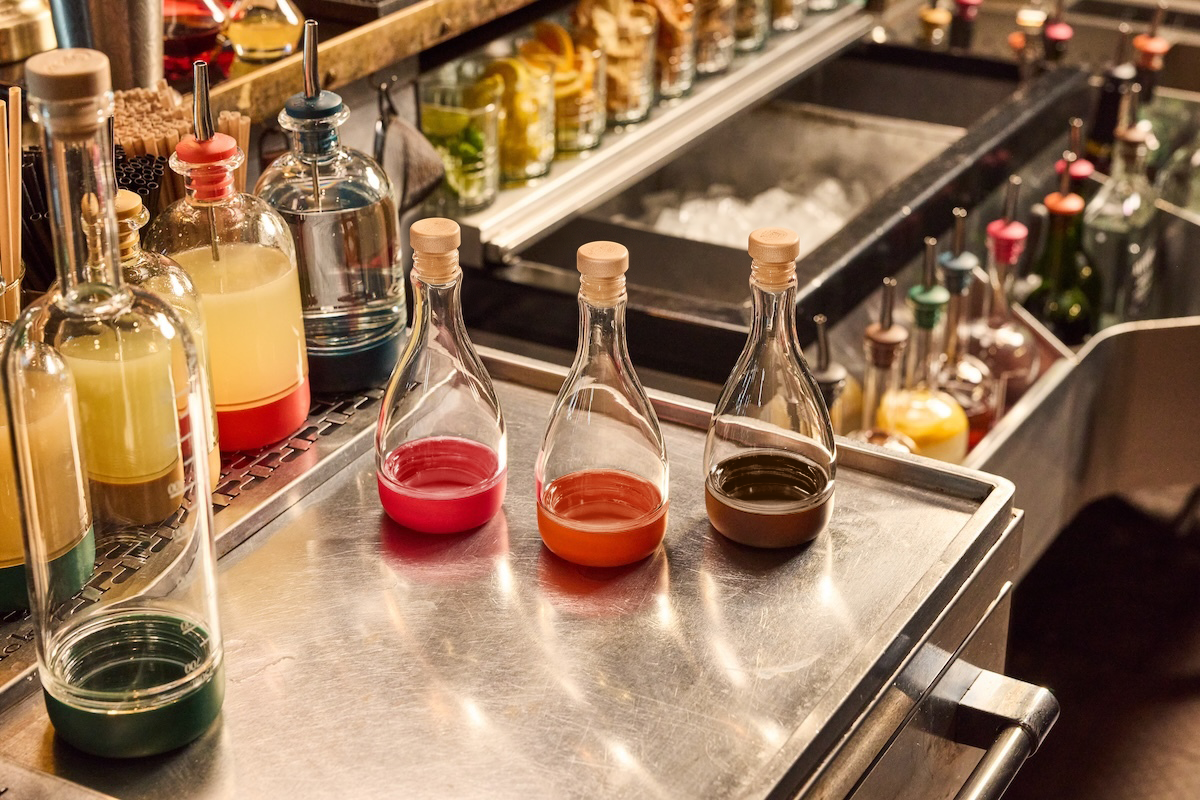 Three small glass bitters bottles with cork stoppers and a screw off bottom, arranged on a stainless steel bar counter with other bar bottles in the background.