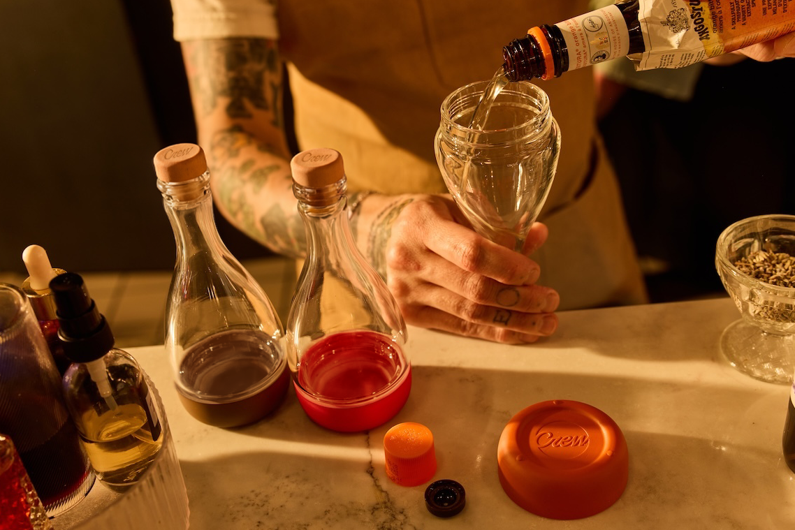 A close-up lifestyle shot of a bartender pouring Angostura bitters into the bottom of a glass bitters bottle that has a screw off bottom, with other glass bitters bottles and cocktail mixing tools on a marble bar counter.