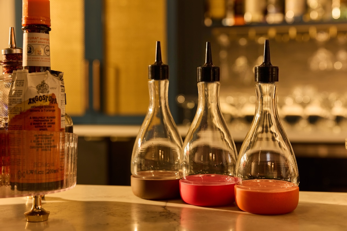Three clear glass bitters dasher bottles with black dasher caps sit on a bar counter, with a removable base next to an Angostura bitters bottle on the left. 