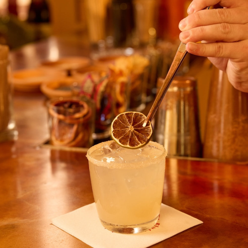 A hand garnishes a pale yellow cocktail in a rocks glass with a dried lime slice, set on a white napkin atop a wooden bar counter.