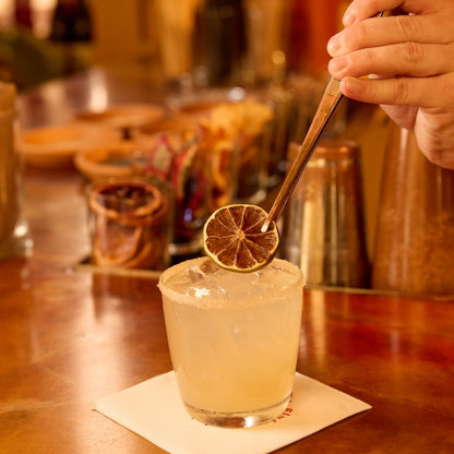A hand garnishes a pale yellow cocktail in a rocks glass with a dried lime slice, set on a white napkin atop a wooden bar counter.