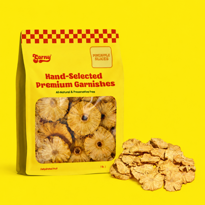 A yellow stand-up pouch labeled 'Carny Hand-Selected Premium Garnishes' with pineapple slices visible through a clear window, shown on a bright yellow background with a pile of dried pineapple slices beside it.