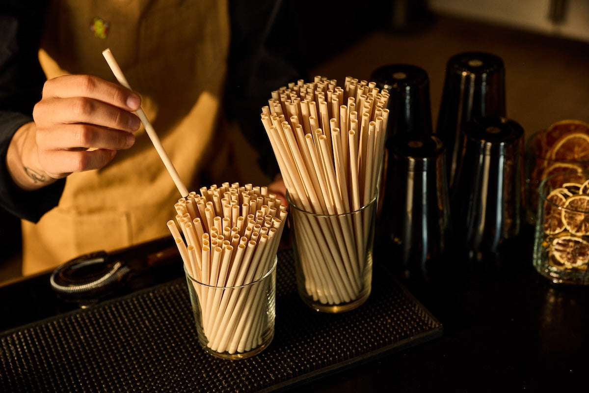 Two clear glass cups filled with compostable plant based cocktail straws on a dark bar counter, with a bartender's hand holding one straw in a dimly lit bar setting.